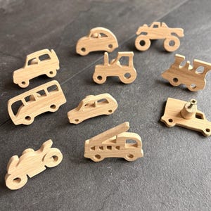Peut inclure: Une collection de petits véhicules jouets en bois, dont une voiture, un bus, un tracteur, un train, un camion, une moto et un camion de pompiers. Les jouets sont en bois clair et sont disposés sur une surface sombre.