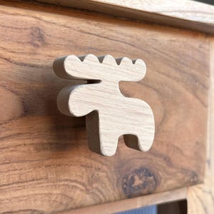 Puede incluir: Tirador de cajón de madera con forma de alce, montado en un cajón de madera. El tirador con forma de alce es de color claro, con una superficie texturizada. El cajón es de madera oscura con vetas visibles.
