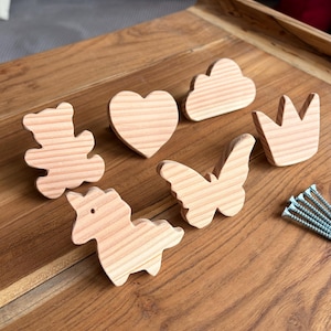 Puede incluir: Una colección de formas de madera sin terminar, que incluye un oso de peluche, un corazón, una nube, una corona, una mariposa y un unicornio. Las formas son de madera clara y están dispuestas sobre una superficie de madera. Se ve un pequeño montón de tornillos.