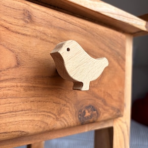 Könnte beinhalten: Ein Nachttisch aus Holz mit einem hellfarbigen, vogelförmigen Schubladengriff. Der Tisch ist aus warm getöntem Holz mit sichtbarer Maserung gefertigt. Der Vogelgriff verleiht dem Möbelstück einen Hauch von Laune. Der Tisch hat eine Schublade und ein Bein.