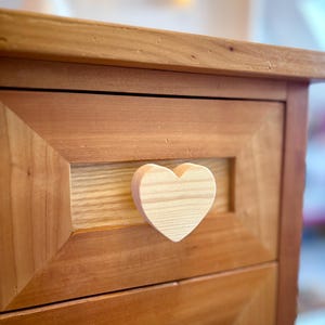 Peut inclure: Un meuble en bois à deux tiroirs, avec des poignées en forme de cœur. Le meuble est en bois brun clair avec une finition naturelle. Les façades des tiroirs présentent un motif de grain de bois contrasté.