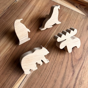 Könnte beinhalten: Vier Holztiere: ein Pinguin, eine Robbe, ein Bär und ein Elch. Jede Figur ist aus hellfarbenem Holz geschnitzt, das die natürliche Maserung zeigt. Die Tiere sind auf einer Holzoberfläche angeordnet.