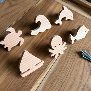 Könnte beinhalten: Eine Sammlung von Holzformen zum Thema Meer, darunter eine Schildkröte, eine Welle, ein Delfin, ein Fisch, ein Oktopus und ein Segelboot. Die Formen sind hell und scheinen unfertig zu sein. Ein kleiner Haufen Schrauben ist ebenfalls sichtbar.