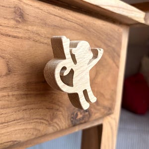 Könnte beinhalten: Holzschubladengriff in Form einer stilisierten Figur, befestigt an einer Holzschublade. Die Figur ist hell und scheint aus einem einzigen Holzstück geschnitzt zu sein. Die Schublade und die umliegenden Möbel sind aus warm getöntem Holz.