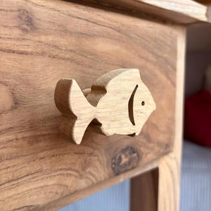 Könnte beinhalten: Eine Holzschublade mit einem fischförmigen Griff. Die Schublade besteht aus hellbraunem Holz mit natürlicher Maserung. Der Fischgriff ist einfach gestaltet und hat ein kleines schwarzes Auge. Die Schublade ist Teil eines größeren Holzmöbels.