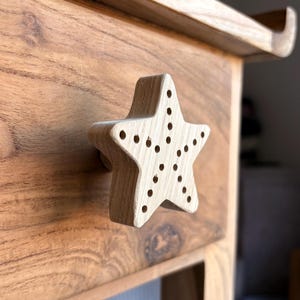 Könnte beinhalten: Ein sternförmiger Holzschubladengriff mit Bohrlöchern, montiert an einer Holzschublade. Schublade und Griff sind aus hellem Holz gefertigt. Der Stern hat ein dreidimensionales Design und ist mit einem kleinen Holzstift befestigt.