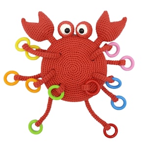Puede incluir: Un juguete de cangrejo de crochet rojo con anillos coloridos unidos a sus patas. El cangrejo tiene ojos saltones y una expresión sonriente.