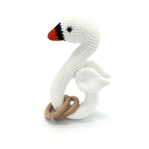 以下が含まれることがあります： 木製の輪が付いた、白の編みぐるみ白鳥のガラガラおもちゃ。白鳥は赤いクチバシと黒い目があります。