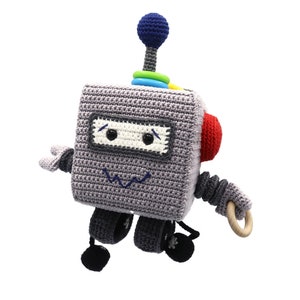 Puede incluir: Un juguete robot de crochet gris con una antena azul, una oreja roja y un anillo de madera. El robot tiene una cara con ojos negros y una sonrisa cosida.