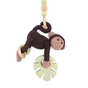 Op de afbeelding: Een bruin gehaakt aapje dat hangt aan een houten ring met een groene, blaadjesvormige bijtring. De aap heeft een bruin lichaam en een lichtbruin gezicht. Het speelgoed is bevestigd aan een groene en houten kralenketting.