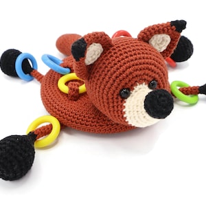 Op de afbeelding: Een gehaakt vosje met zwarte en witte gezichtskenmerken en een bruin lichaam. Het speelgoed heeft vier zwarte en witte gehaakte armen met kleurrijke plastic ringen.