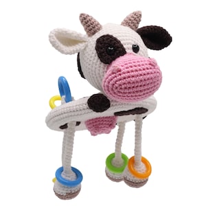 Puede incluir: Un juguete de vaca de crochet con manchas blancas y negras y una nariz rosa. El juguete tiene tres patas con anillos coloridos unidos a ellas.