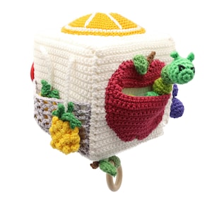 May include: A crocheted cube toy with different textures and colours. It features a red apple with a green worm inside, a yellow pineapple, and a purple grape. The cube has a wooden ring attached to it.