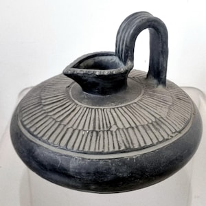 Puede incluir: Un recipiente de cerámica gris oscuro, de estilo antiguo, con tapa y asa. La tapa presenta un patrón radial, y el asa se arquea con gracia. El pico es pequeño y detallado. La pieza tiene un aspecto envejecido, lo que sugiere edad y uso.