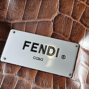 Könnte beinhalten: Nahaufnahme einer braunen Lederoberfläche mit Krokodilhautstruktur. Eine silberne rechteckige Platte ist angebracht, auf der der Markenname "FENDI" in schwarzen Großbuchstaben mit "casa" darunter steht. Das eingetragene Warenzeichen ist sichtbar.
