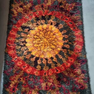 May include: A rectangular shag rug with a vibrant, abstract design. The rug features a central burst of orange and yellow, encircled by rings of red, black, and brown. The outer edges are a muted gray-green, creating a striking contrast.