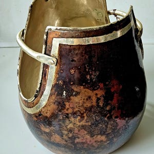 Op de afbeelding: Een handgemaakt, decoratief vat met een unieke vorm. De binnenkant is goudkleurig, terwijl de buitenkant een gevlekte, donkerbruine en koperen afwerking heeft. Zilverkleurige metalen accenten sieren de rand en de handgreep.