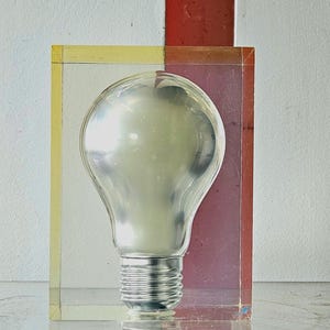 Könnte beinhalten: Eine klare Glühbirne, die in einem transparenten rechteckigen Block eingeschlossen ist. Der Block hat auf einer Seite einen gelben Rand und auf der anderen Seite einen rot-violetten Abschnitt. Die Glühbirne hat einen silbernen Sockel und ist im Block zentriert.
