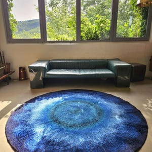 May include: A round, shaggy rug with a gradient of blues, from deep navy to light turquoise. The rug is centered in a room with a green leather sofa and large windows overlooking a lush, green landscape.