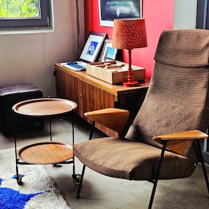 Könnte beinhalten: Ein Wohnzimmer im Retro-Stil mit einem braunen Sessel, einem zweistöckigen Beistelltisch und einem gemusterten Teppich. Eine rote Lampe und ein Vintage-Filmplakat ergänzen die Szene.