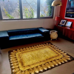 May include: A rectangular shag rug with a yellow center and a border of brown and gold leaf-like designs. The rug is placed on a light-colored floor in a room with a black leather sofa and a large window.
