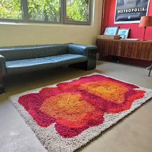 Könnte beinhalten: Ein Shaggy-Teppich mit einem lebendigen Blumenmuster in Rot-, Orange- und Rosatönen. Der Teppich hat einen neutralfarbenen Rand und liegt auf einem Betonboden. Ein Blickfang für jeden Wohnraum.