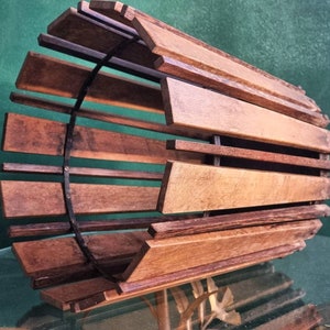 Könnte beinhalten: Ein Lampenschirm aus Holz mit zylindrischer Form und einer dunkelbraunen Oberfläche. Der Schirm besteht aus dünnen Holzlatten, die in einem kreisförmigen Muster angeordnet sind. Der Schirm hat einen Metallrahmen, der an den oberen und unteren Rändern sichtbar ist.