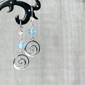 May include: A pair of silver earrings with a spiral design. Each earring features a light blue bead and a smaller star-shaped accent. The earrings are displayed on a black stand against a neutral background.