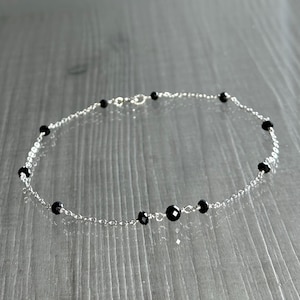 May include: A delicate silver anklet featuring small, faceted black beads evenly spaced along a fine chain. The anklet has a simple clasp closure and is displayed on a light gray surface.