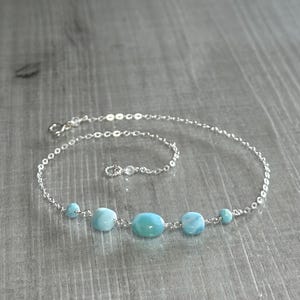 May include: A delicate silver chain necklace featuring five light blue, oval-shaped gemstones. The necklace has a small clasp and is displayed on a wooden surface. The gemstones are spaced evenly along the chain.