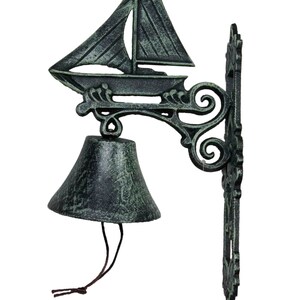 May include: A green cast iron door bell with a sailboat design and a bell hanging from the bottom. The bell has a brown rope attached to it.