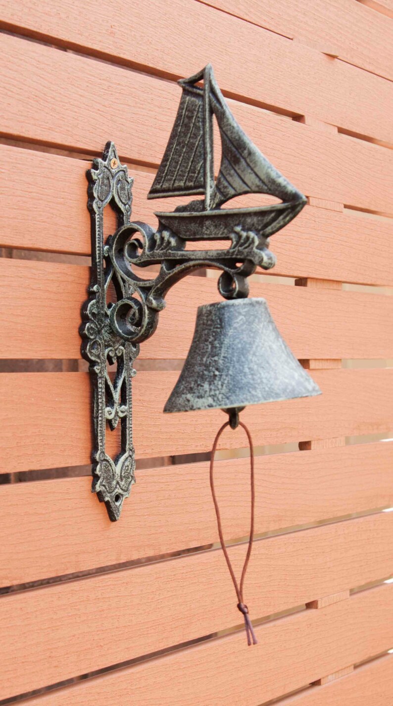 May include: A cast iron door knocker with a sailboat design and a bell. The knocker is mounted on a wooden wall with a brown stain.