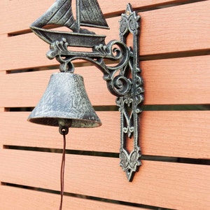 May include: A black cast iron door knocker with a sailboat design and a bell. The knocker is mounted on a wooden door.