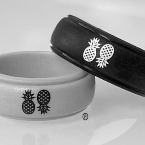 May include: Two silicone rings, one light grey with a black pineapple graphic and one black with a white pineapple graphic. Each ring features a registered trademark symbol near the bottom edge, showcasing a smooth, rounded band design.