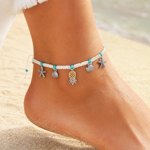 Op de afbeelding: Witte macramé enkelband met turquoise kralen en bedels. Bedels zijn onder meer een gouden ananas, zilveren zeesterren en schelpenhangers. De enkelband wordt gedragen op een voet op een zandstrand.
