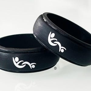 Pode incluir: Dois anéis de silicone preto com um gráfico branco de duas figuras estilizadas ligadas numa posição reclinada. Os anéis têm um acabamento mate liso com um detalhe de banda central elevada e um pequeno símbolo de marca registada.