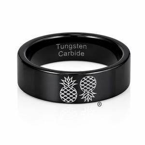 Puede incluir: Un anillo negro de carburo de tungsteno con dos iconos de piña grabados en la banda exterior. El interior de la banda tiene la inscripción Tungsten Carbide. El diseño incluye un pequeño símbolo de marca registrada cerca de las piñas.