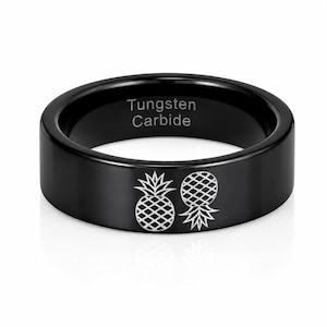 Puede incluir: Un anillo de carburo de tungsteno negro con acabado pulido y dos iconos de piña grabados en la banda exterior. El interior de la banda está claramente marcado con el texto Tungsten Carbide, indicando la composición del material del anillo.