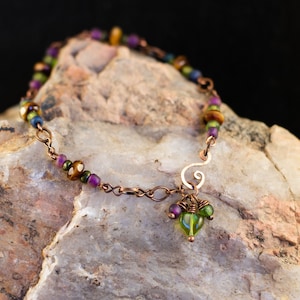 Könnte beinhalten: Ein Armband aus Kupferkette mit einer Vielzahl von bunten Perlen, darunter lila, grün, braun und gold. Das Armband hat einen Anhänger mit einer grünen herzförmigen Perle und kleineren grünen Perlen.