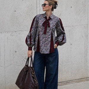 May include: A patterned blouse with burgundy panels and a bow detail. The blouse features long, puffed sleeves and is worn with dark denim jeans. A large, brown leather tote bag is carried.