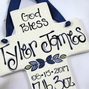 May include: White ceramic cross with blue and green painted leaves and the text "God Bless Tyler James 08.15.2017 7lb 3oz".