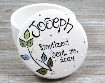 May include: A round white ceramic keepsake box with a removable lid. The lid features hand-painted black text reading Joseph Baptized Sept. 28, 2024, accented by a decorative vine with green and blue leaves and small white dots.