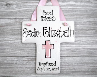 May include: White ceramic cross with pink accents. The cross has the text "God Bless Sadie Elizabeth Baptized Sept. 22, 2024" written on it.
