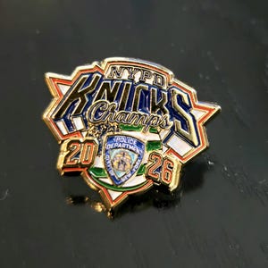 Puede incluir: Un pin de solapa de metal dorado con el texto NYPD KNICKS Champs en letras azules y doradas. El diseño incluye un emblema central en forma de escudo del Departamento de Policía de Nueva York, con los números 20 y 26 en los lados.