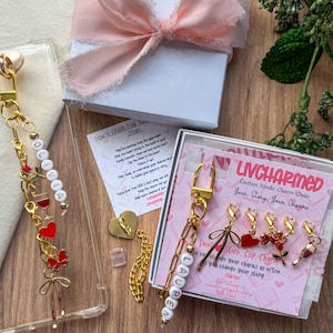 May include: A collection of custom charm chains with gold-tone hardware, featuring personalized names and decorative charms like hearts, butterflies, and wine glasses. The chains are displayed with a gift box tied with a pink ribbon and a card that says "LIVCHARMED".