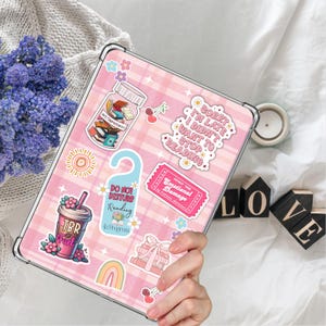 May include: A pink and white checkered iPad case with various stickers, including a jar of books, a do not disturb sign, a ticket that says "Adult Only Emotional Damage", a rainbow, and a cup of coffee with the text "TBR Fuel".