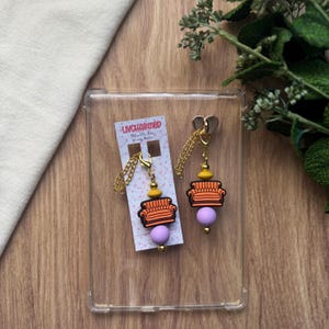 May include: A pair of gold-colored chain earrings with a miniature orange couch charm, yellow bead, and a purple bead. The earrings are displayed on a clear plastic case with a tag that reads "UNCHARMED".