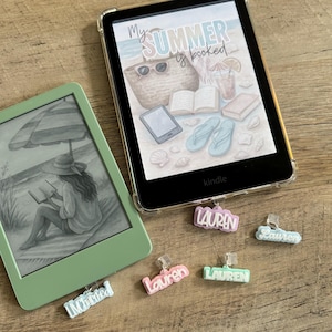 May include: Two e-readers, one mint green and one black, with decorative charms. The black e-reader displays the text "My Summer is Booked" with beach-themed illustrations. Several charms with the name "Lauren" are attached.