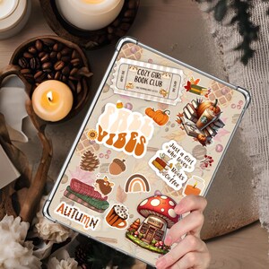 Puede incluir: Una tableta con una funda transparente está cubierta de pegatinas con temática otoñal. Las pegatinas incluyen calabazas, libros, una casa de setas y las palabras "Cozy Girl Book Club", "Vibes", "Autumn" y "Just a Girl who loves Books & Coffee".