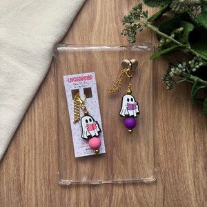 May include: A pair of gold-toned chain earrings featuring a white ghost holding a pink book. The ghost charm is above a pink bead on one earring and a purple bead on the other. The earrings are displayed on a clear case.